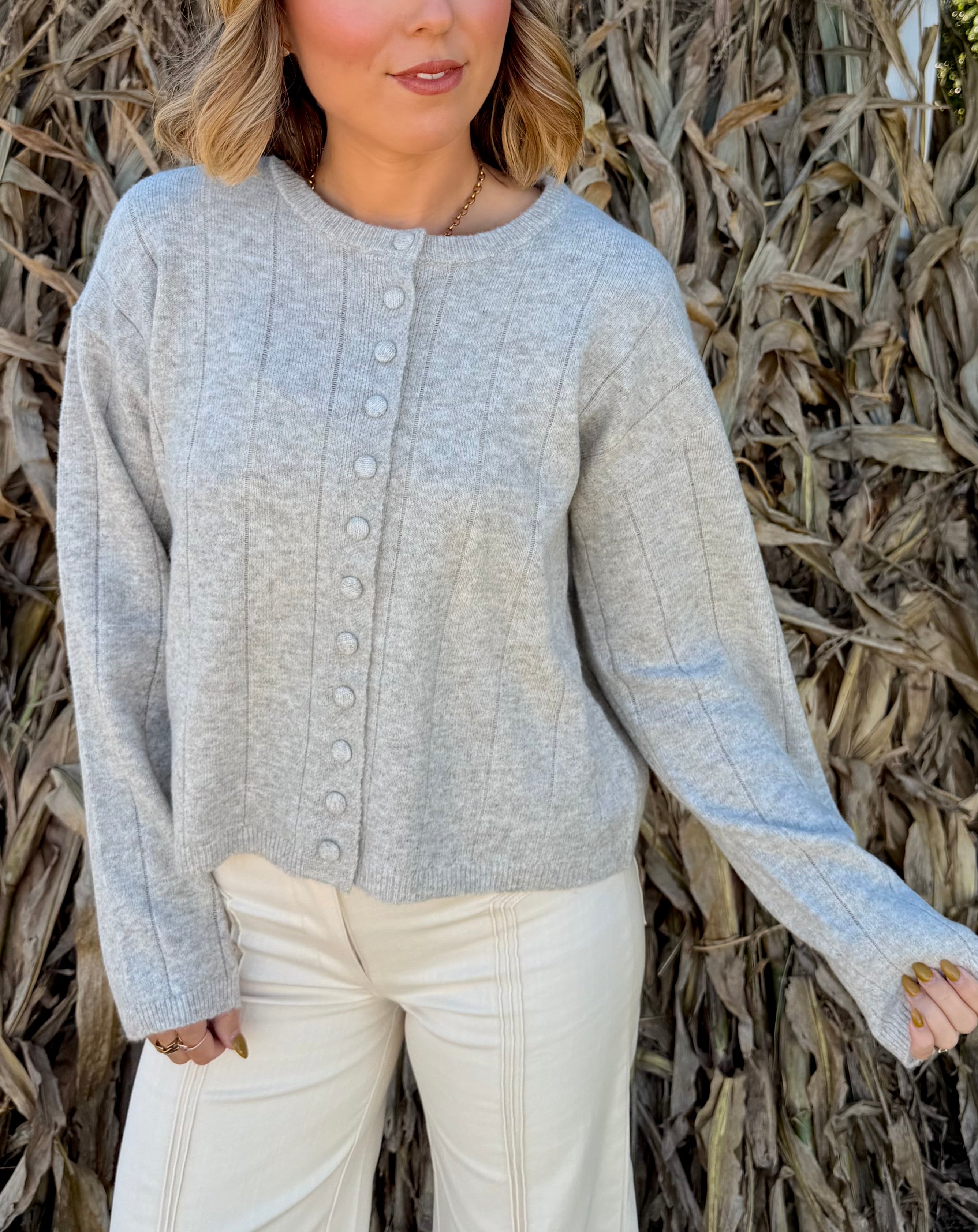 Model wearing a light heather grey knit cardigan with a round neckline, fabric-covered button front, and long sleeves, paired with cream trousers.