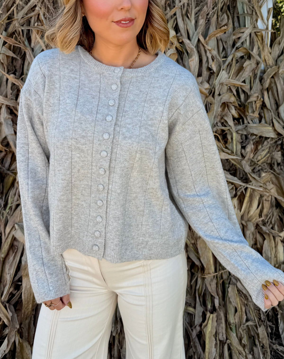 Model wearing a light heather grey knit cardigan with a round neckline, fabric-covered button front, and long sleeves, paired with cream trousers.
