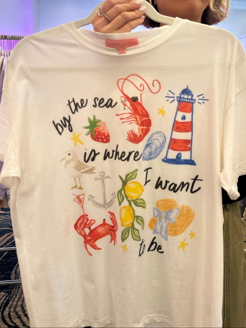 White t-shirt with beach-themed graphics and text on a hanger Person holding a t-shirt with beach-themed graphics and text in a store setting, summer staple piece, fruits and sea animals, beachy details, summer and ocean, graphic tee, straight fit 