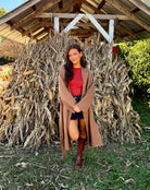 Model wearing a camel double-breasted wrap coat with a waist-cinching tie belt, long sleeves, and a trench-inspired longline silhouette.