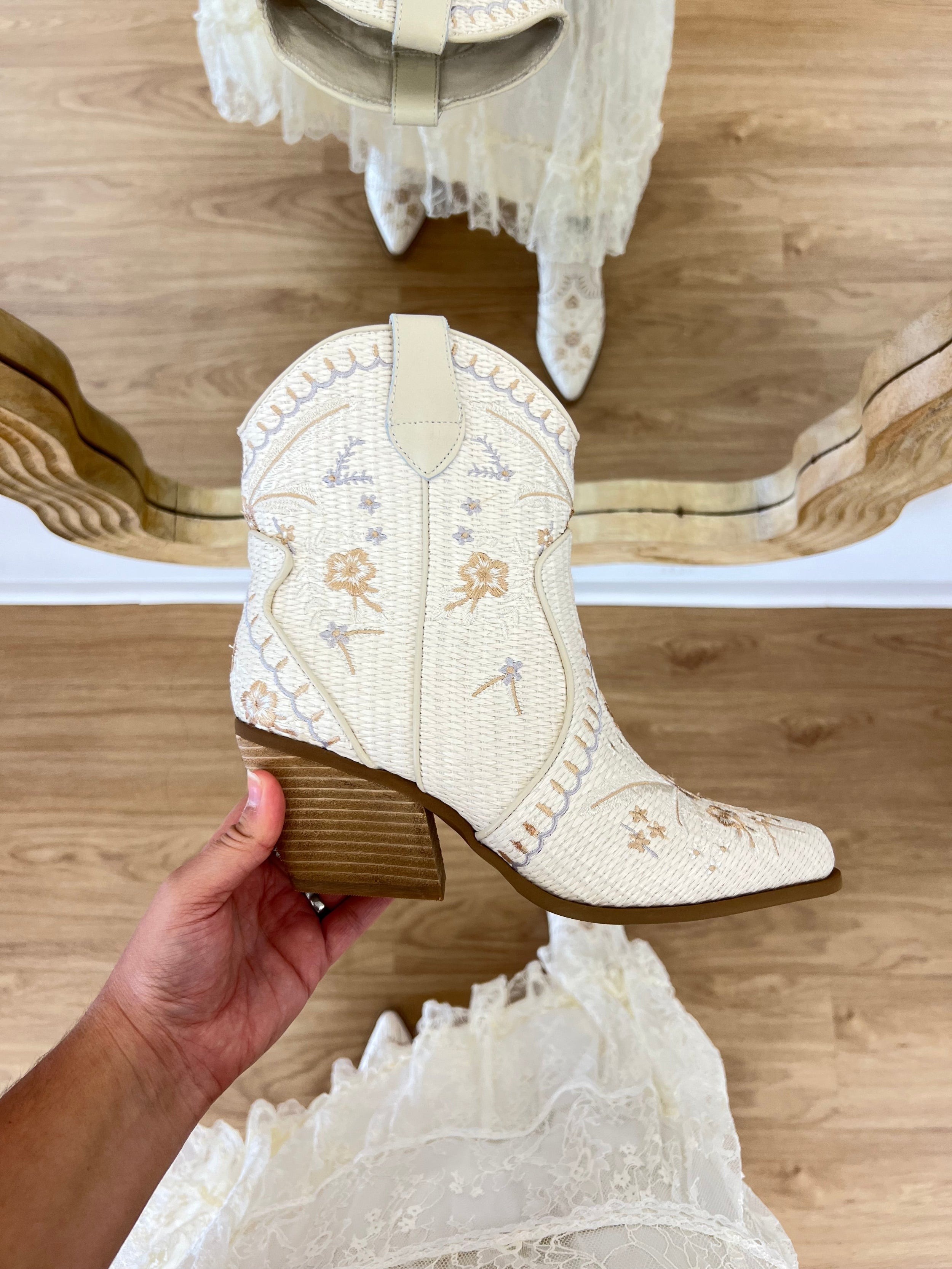 White embroidered boot held by a hand on a wooden floor with lace fabric. coastal cowgirl, raffia, western beach boot