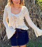 Model wearing a cream pointelle knit V-neck sweater with a cascading ruffle front, ruffled hem, and long bell sleeves, paired with a dark denim mini skirt.