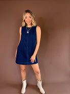 Dark-wash sleeveless denim mini dress with a high round neckline, exposed seams, and a flowy skirt.