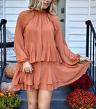 Model wearing the Friendsgiving ruffle mini dress in ginger rust orange with a high neckline, long sleeves, a gathered waist, and a tiered ruffle mini hem.