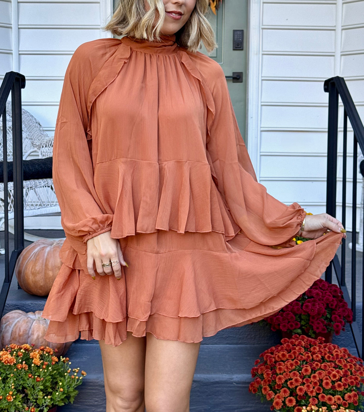 Model wearing the Friendsgiving ruffle mini dress in ginger rust orange with a high neckline, long sleeves, a gathered waist, and a tiered ruffle mini hem.