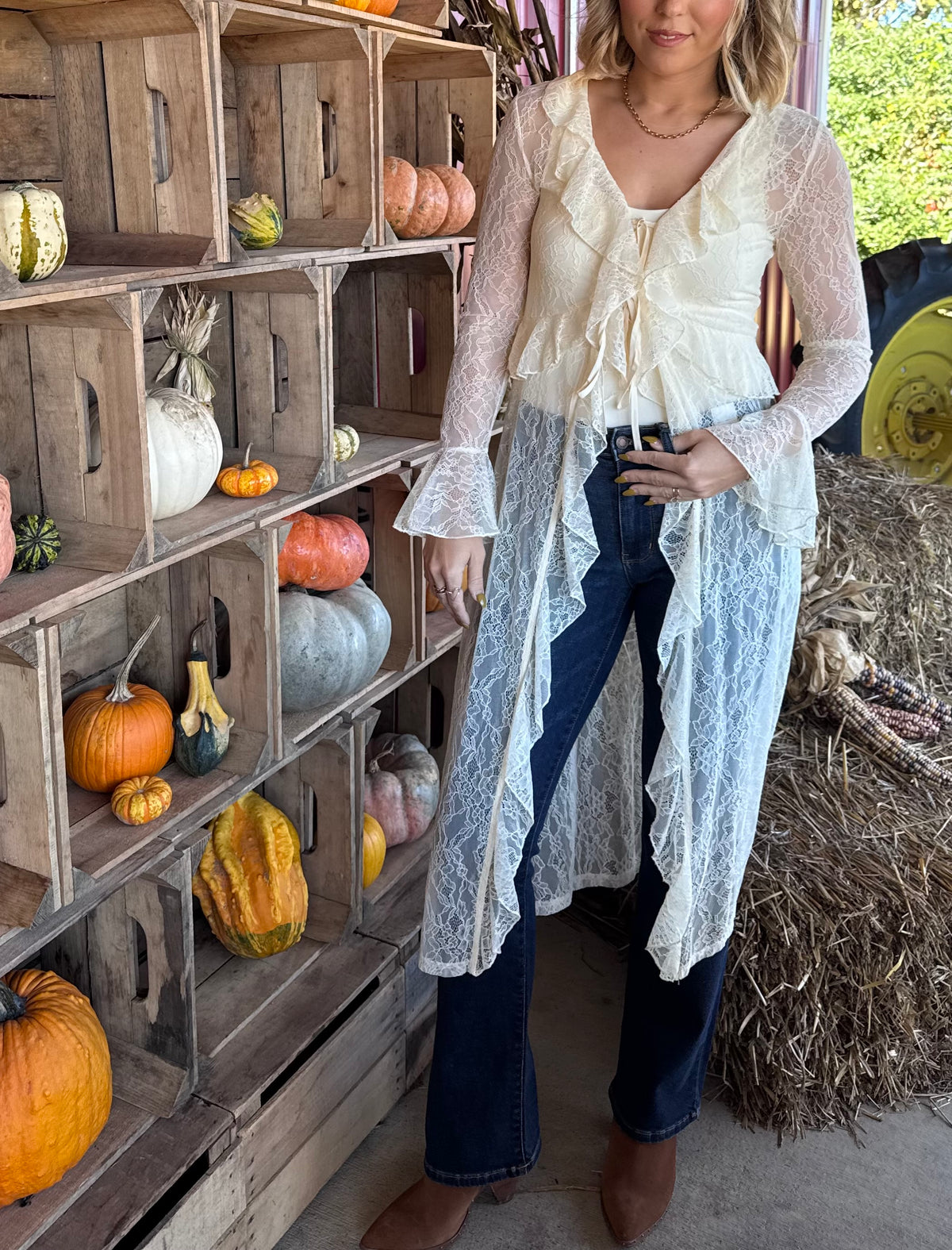 Model wearing an ecru sheer lace duster with cascading ruffle trim, a tie-front closure, and long bell sleeves, layered over a white top and dark flare jeans.