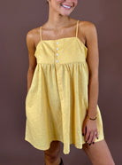 Model wearing the Honey Days honey yellow mini dress with thin straps, a button-detail bodice, and a gathered skirt against a brown backdrop.