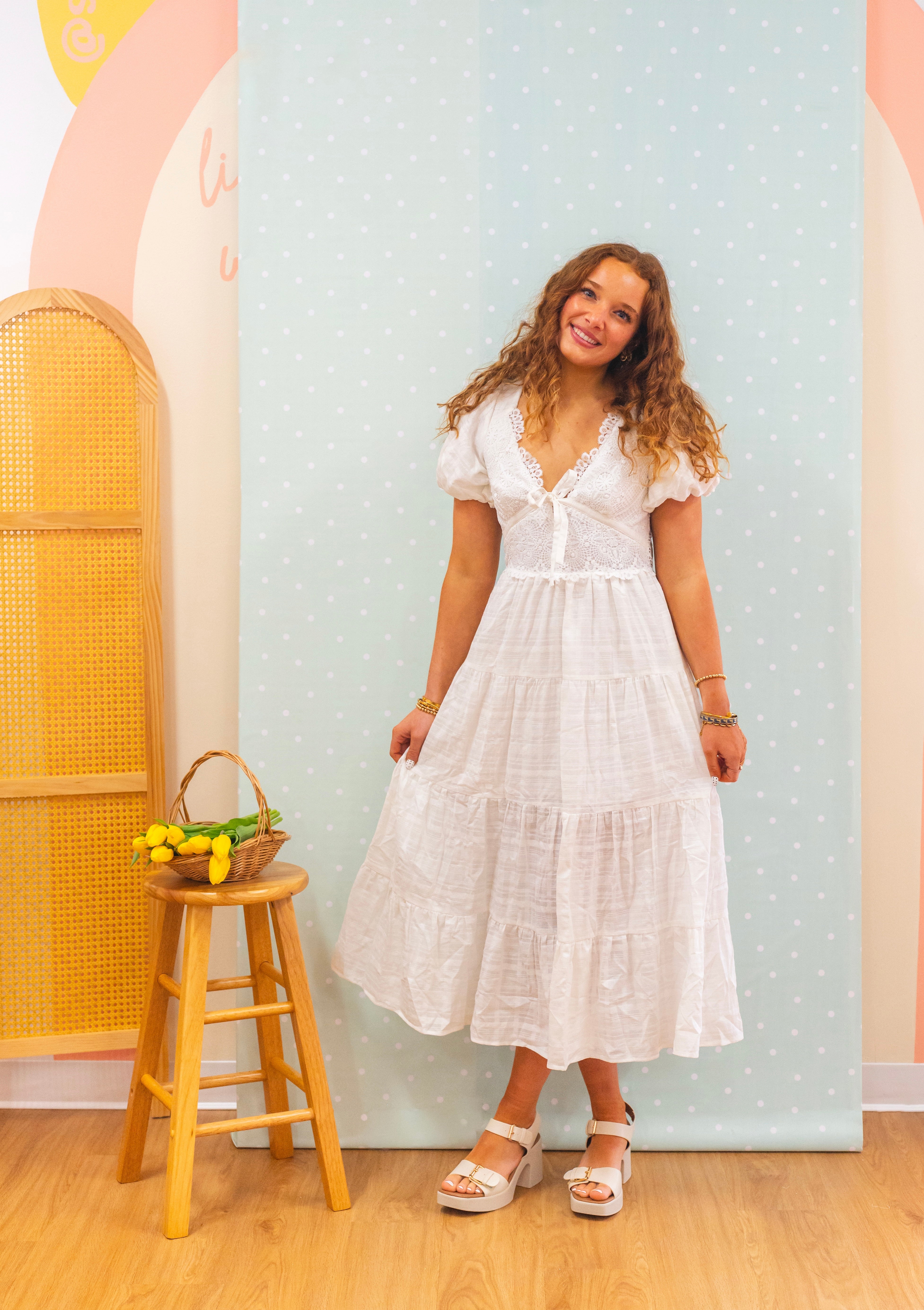 Woman in a white dress standing in a room with a colorful wall and wooden stool, bridal dress, bridal shower, lace dress, maxi dress, a line dress