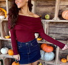 Model wearing a burgundy off-the-shoulder long-sleeve top with a draped fold-over neckline, paired with a dark denim mini skirt.