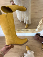 A pair of mustard yellow suede knee-high boots with brown stacked block heels, shown in a mirror reflection and handheld view on a wood floor. A person wearing a white lace dress is visible in the reflection, and a hand holds one boot while another is held up to show the interior.