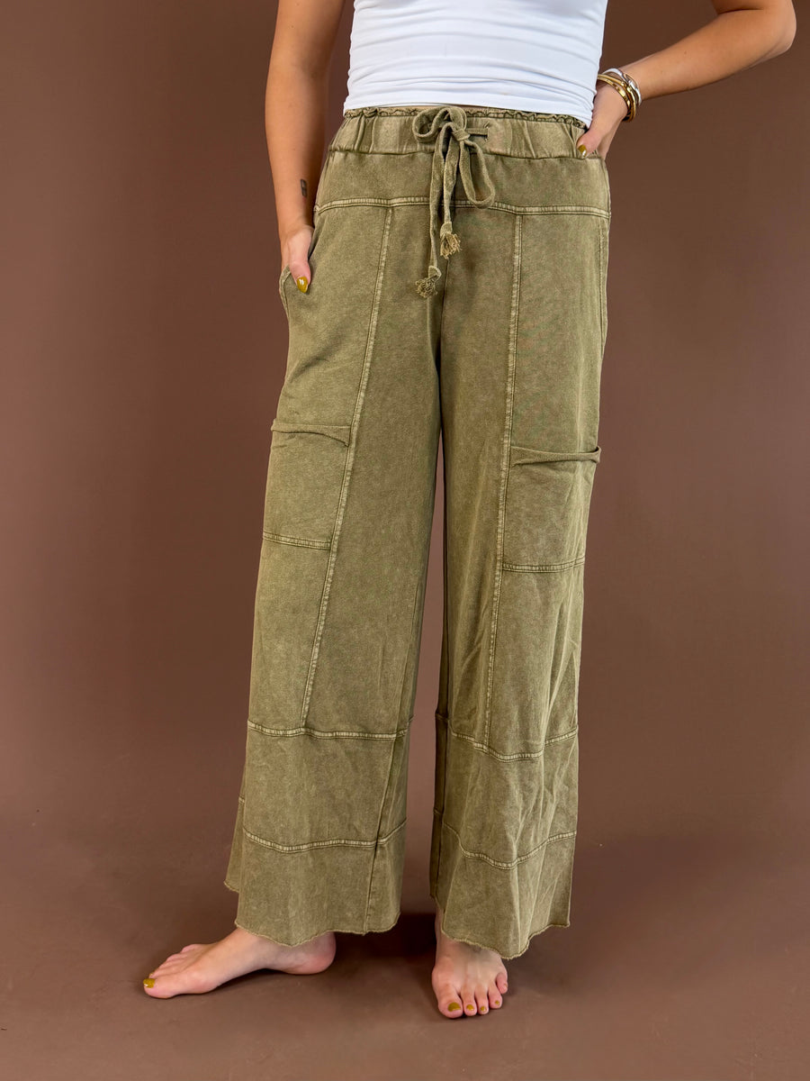 Front view of a model wearing Olive Mist olive mineral-wash wide-leg pants with a drawstring waist, patch pockets, and raw hems.