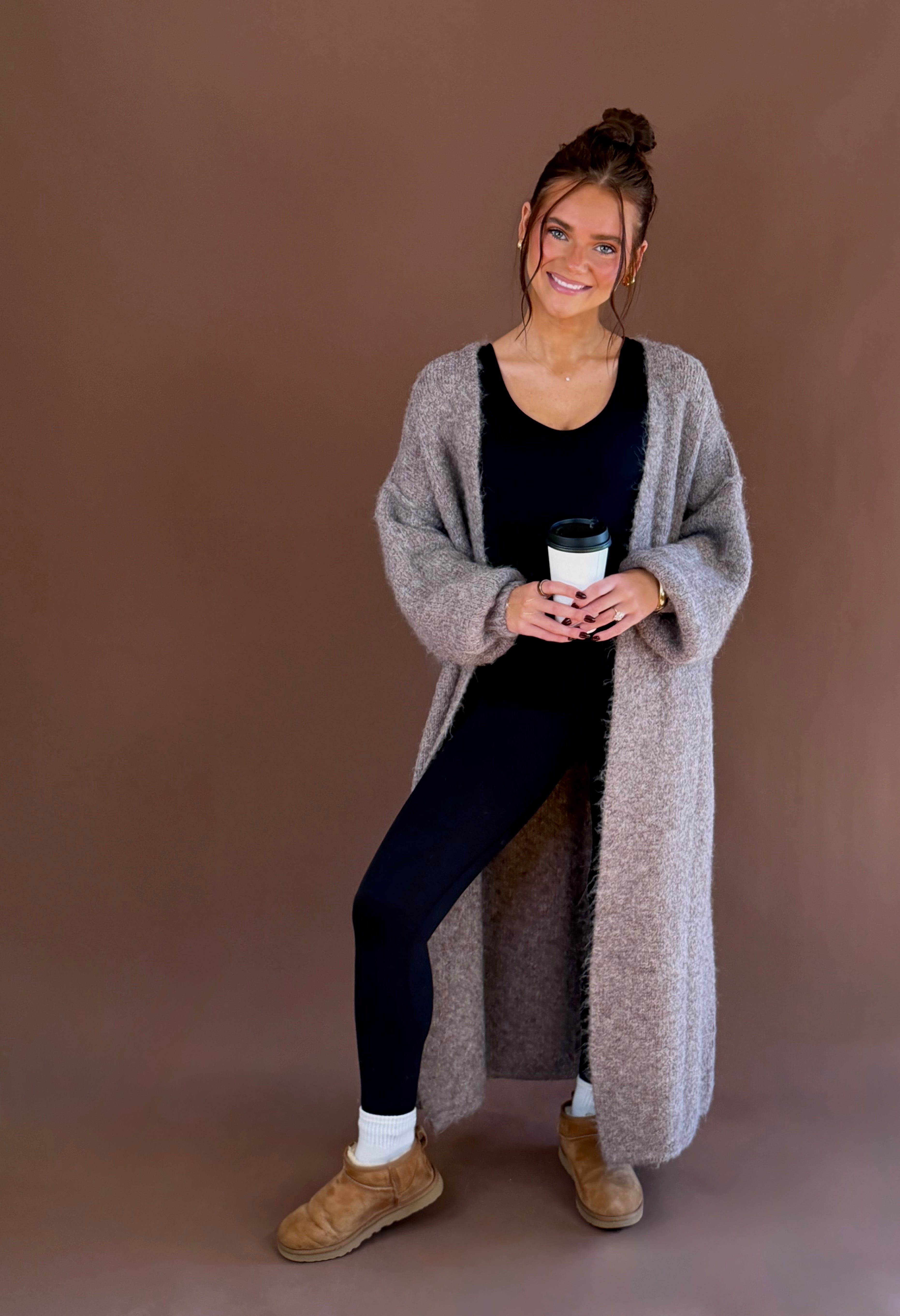 Model wearing an ash chocolate long knit duster cardigan over a black outfit with white socks and brown clogs.