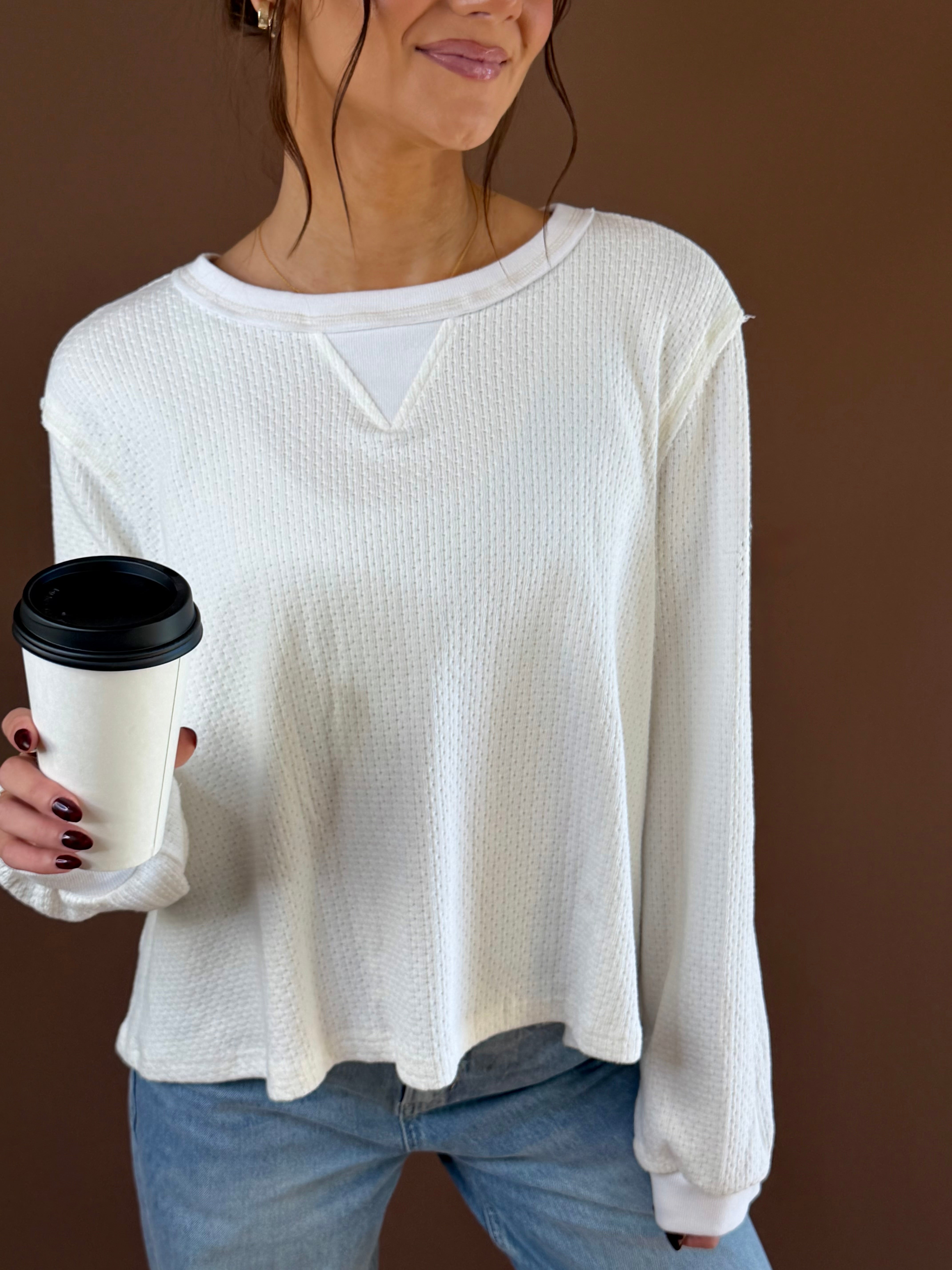 Model wearing an off-white vintage wash long sleeve knit top with ribbed texture and seam details, holding coffee and wearing jeans.