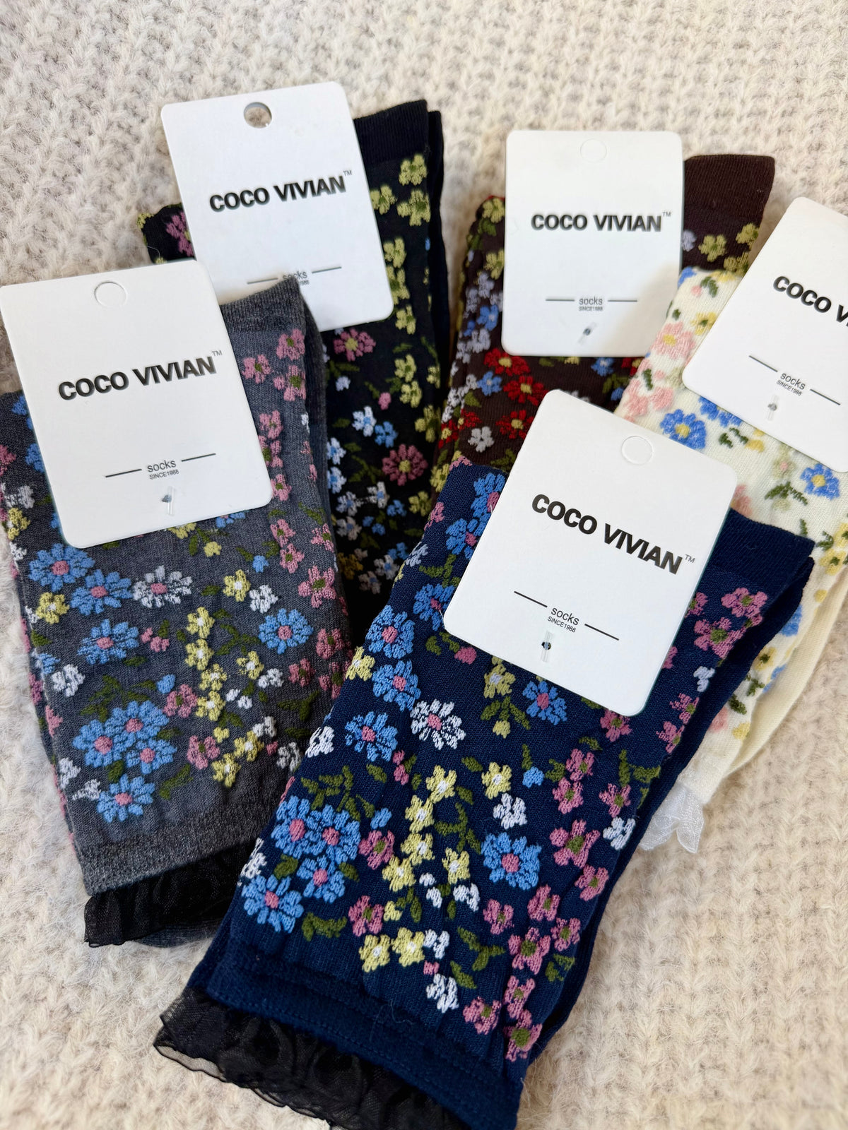 Five pairs of floral crew socks in cream, brown, black, grey, and navy, arranged on a textured cream surface.