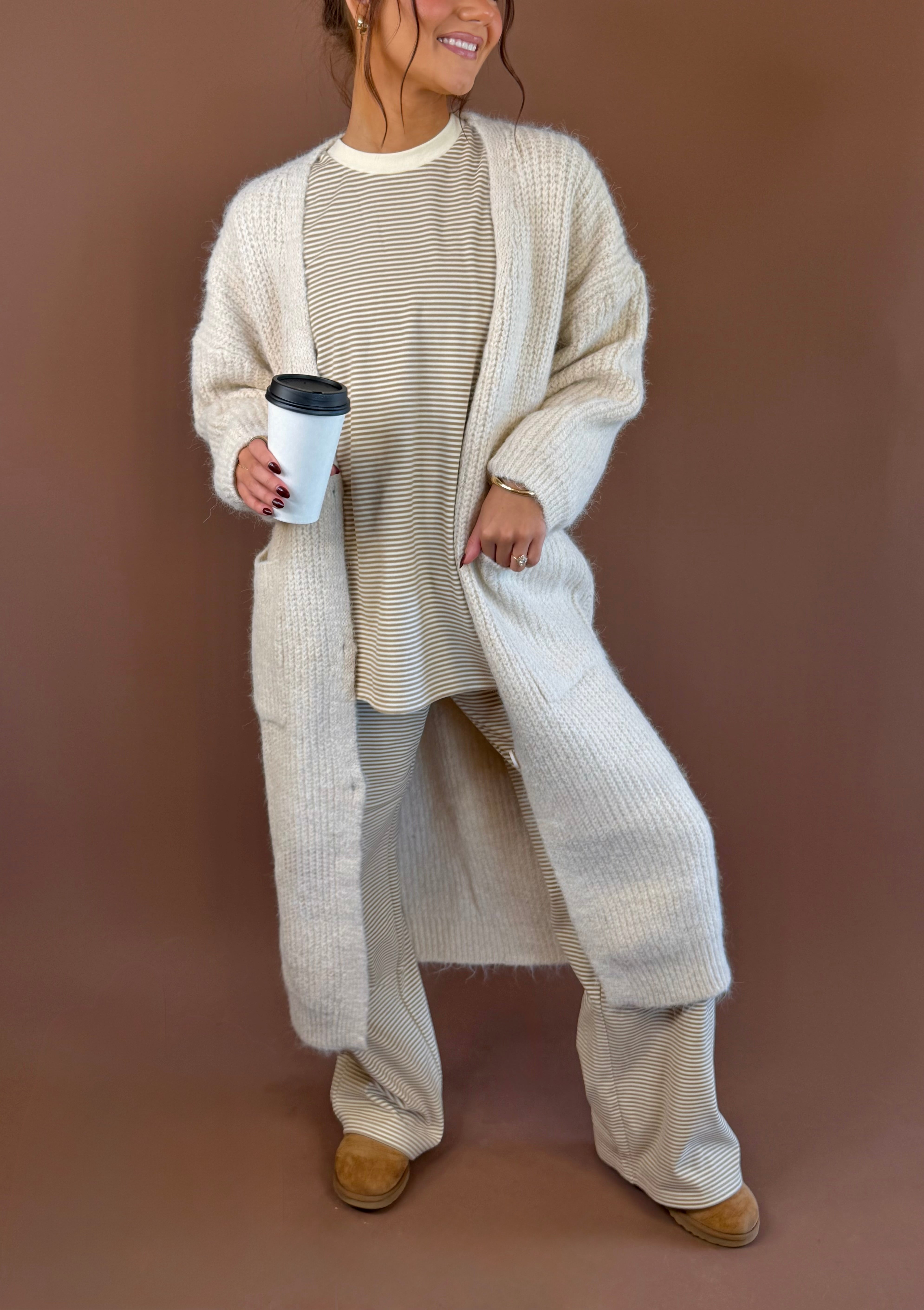 Model wearing an almond chunky knit long cardigan over a light top and lounge pants, holding a coffee cup.