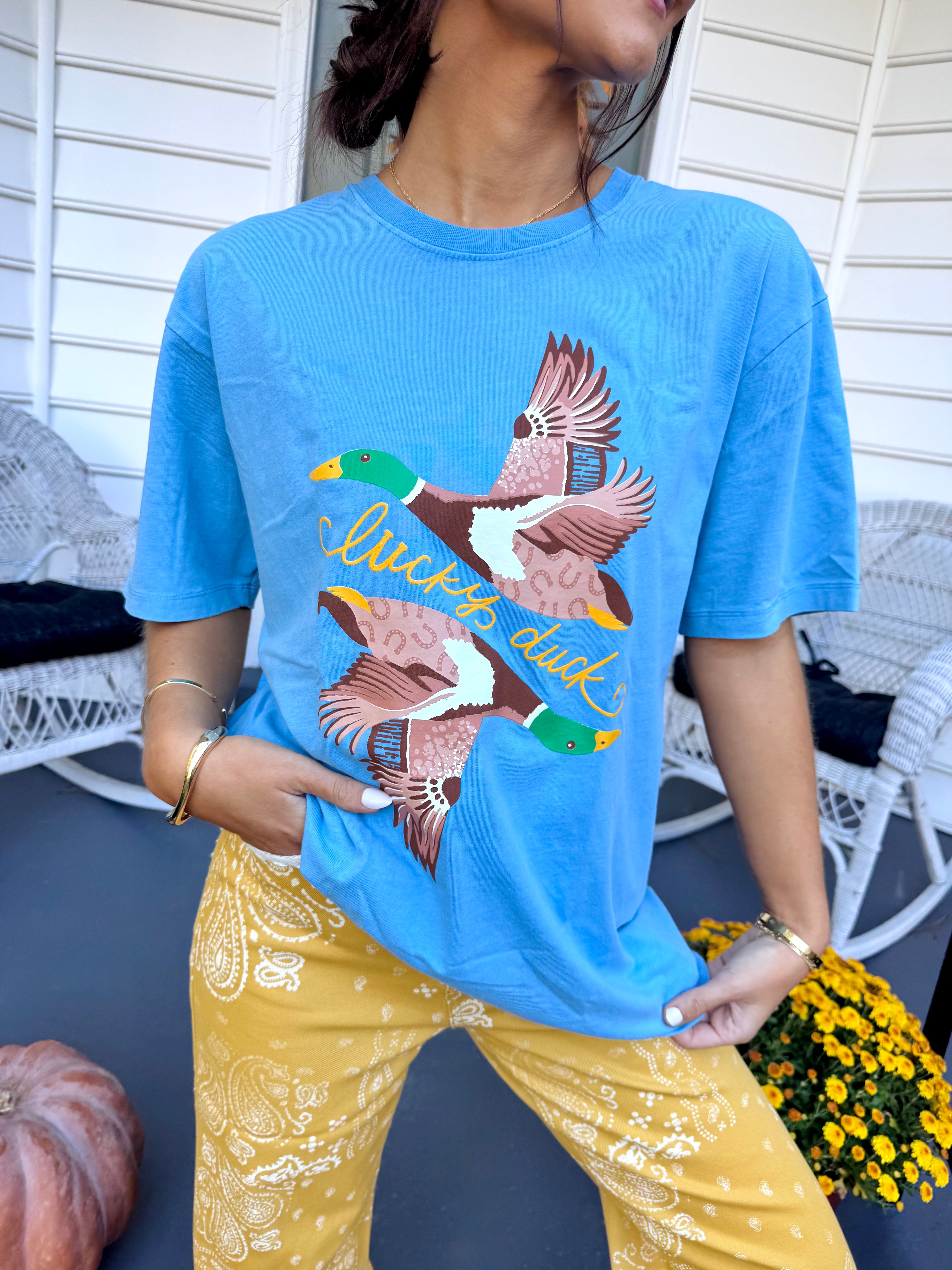 Model wearing a bright blue oversized graphic tee printed with two flying mallard ducks and yellow 'lucky ducks' script, styled with mustard paisley pants.