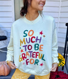 Model wearing a cream crewneck sweatshirt with colorful hand-lettered text that reads SO MUCH TO BE GRATEFUL FOR with star accents.
