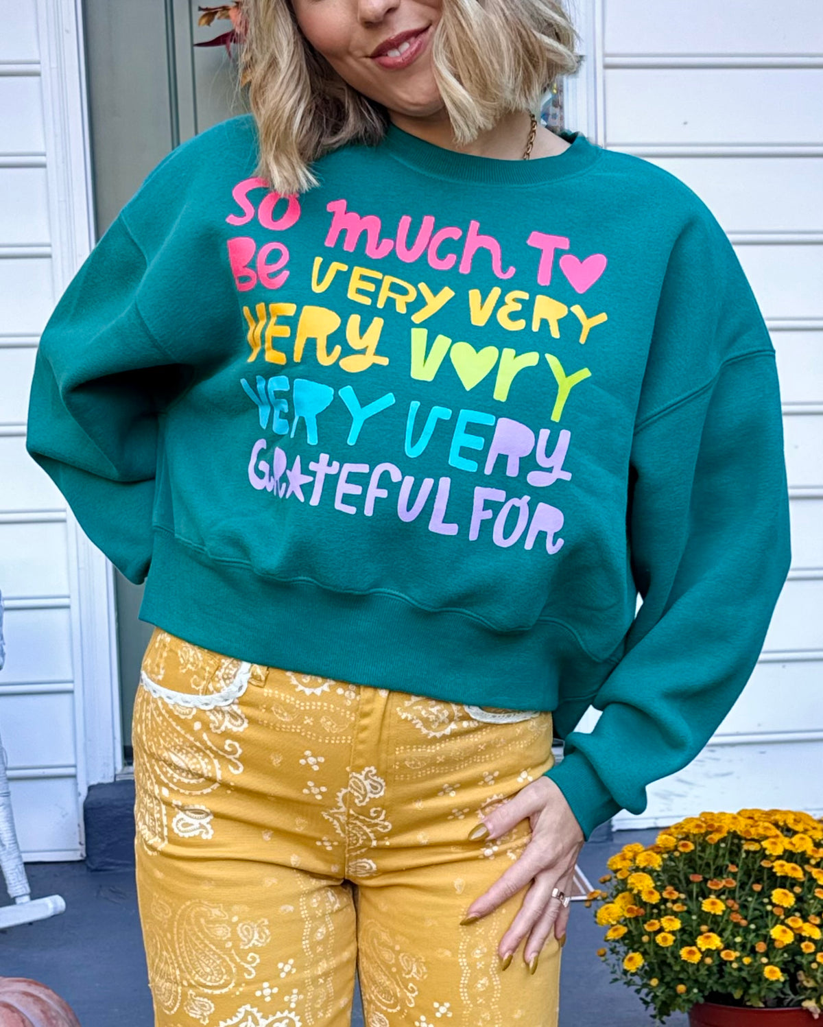 Model wearing a teal green cropped crewneck sweatshirt with colorful lettering that reads SO MUCH TO BE VERY VERY VERY VERY GRATEFUL FOR with heart details.