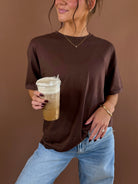Model wearing a brown relaxed boyfriend T-shirt with short sleeves and a round neckline, holding a coffee cup and wearing jeans.