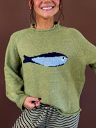 Model wearing an olive green mock neck sweater with a blue sardine graphic across the chest and long sleeves.