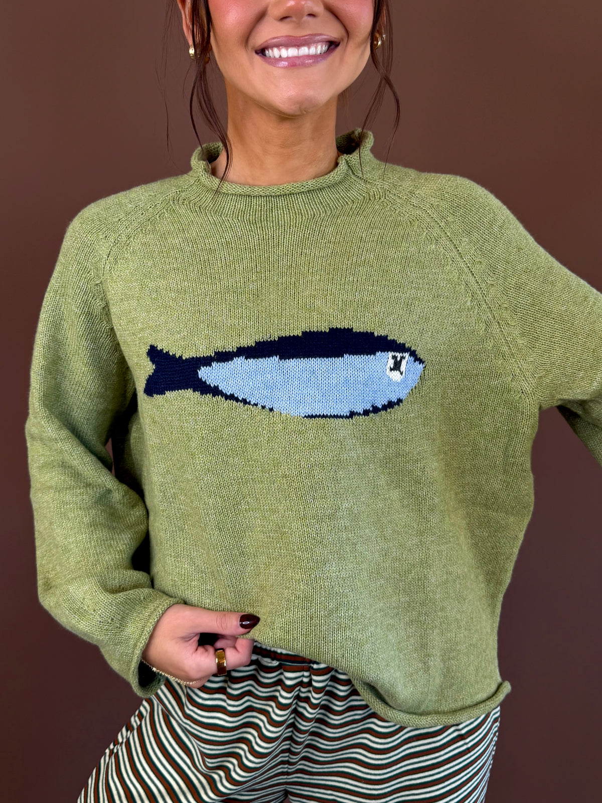 Model wearing an olive green mock neck sweater with a blue sardine graphic across the chest and long sleeves.