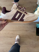 Beige and brown sneaker with decorative elements held by a hand on a wooden floor.