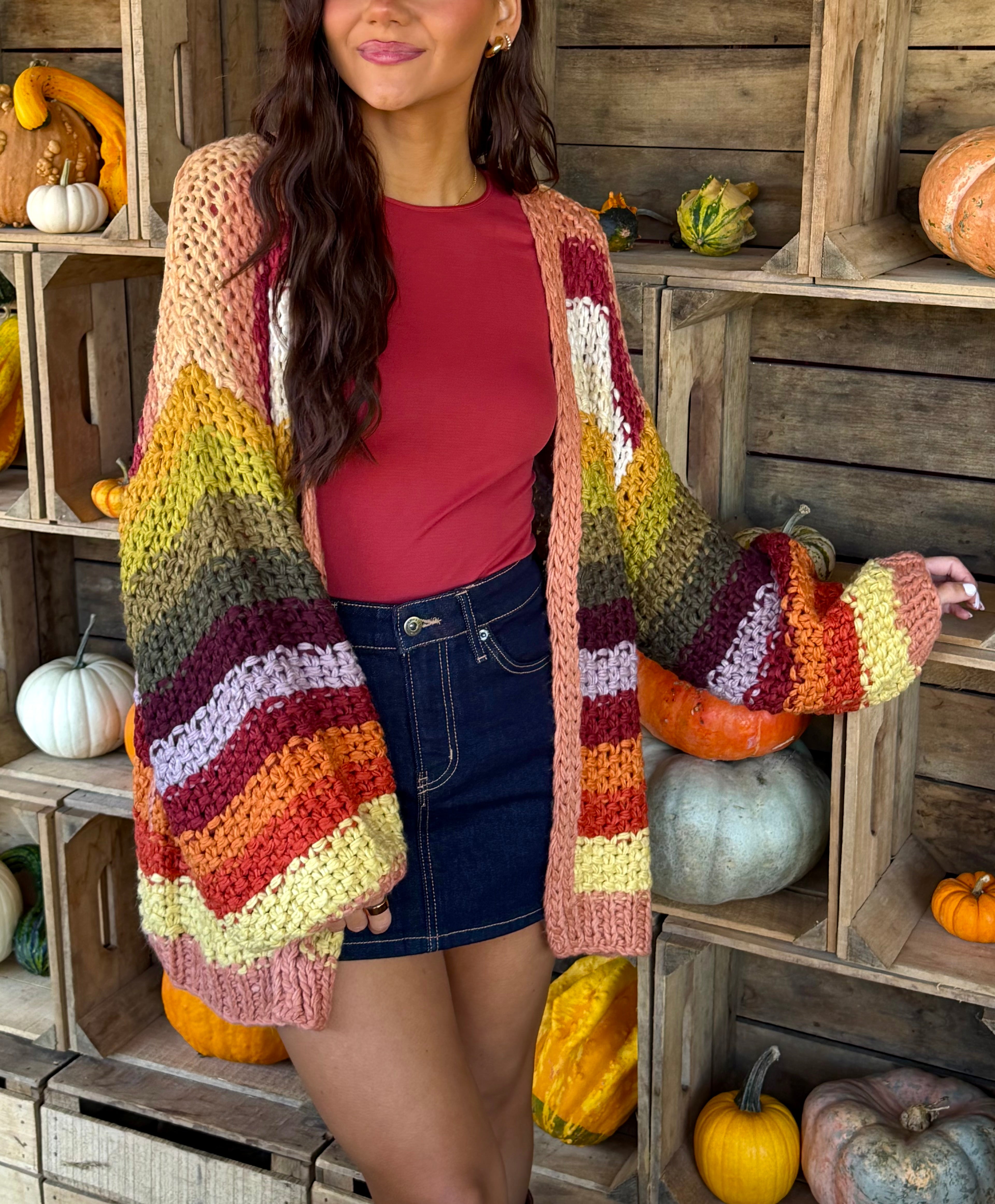 Model wearing a chunky hand-crochet open-front cardigan in autumn-inspired multicolor stripes of pink, mustard, olive, rust, and burgundy.