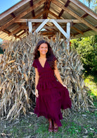 Model wearing a dark wine midi dress with a deep V-neckline, flutter short sleeves, a smocked waist, and a tiered ruffle skirt, styled with brown boots in front of dried cornstalks.