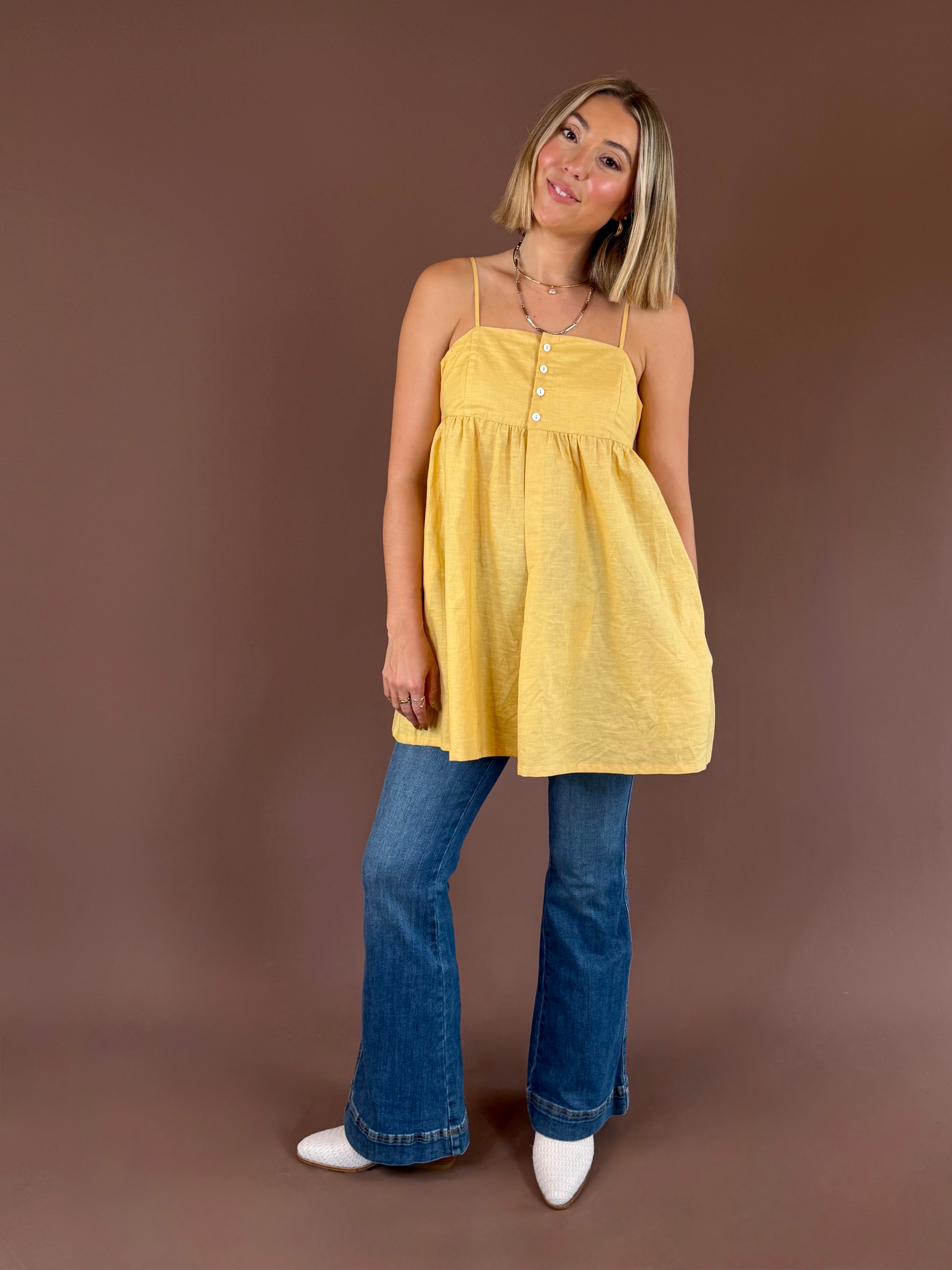 A woman modeling a yellow sleeveless top with thin straps and a buttoned chest detail, paired with blue flared jeans and white pointed shoes, against a solid brown studio backdrop.